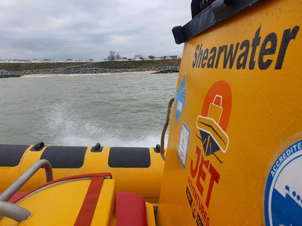 Yellow inflatable boat "Shearwater" moving on water by rocky shore under cloudy sky.