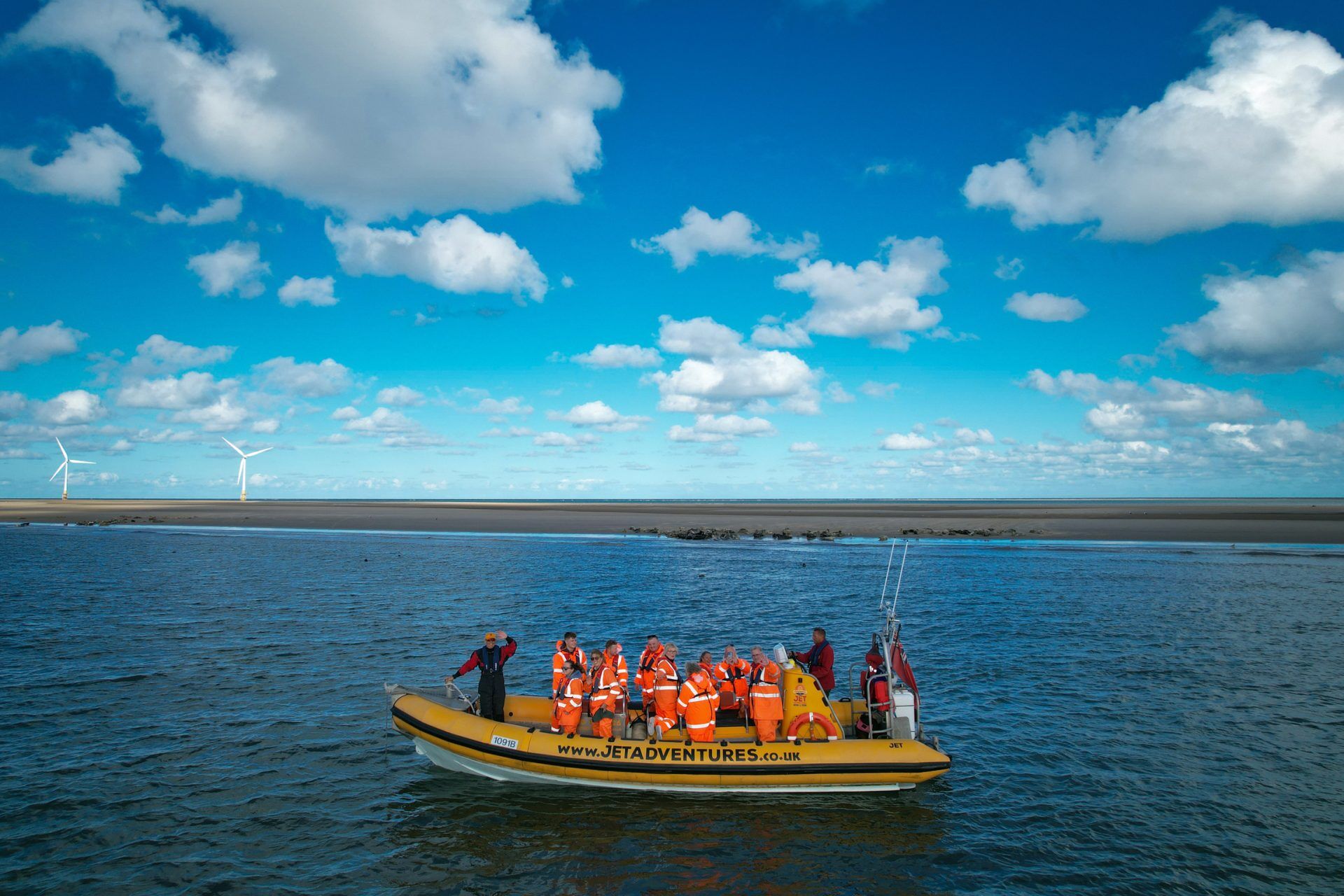 People in orange safety gear on a yellow boat, sandy shore with wind turbines in background.