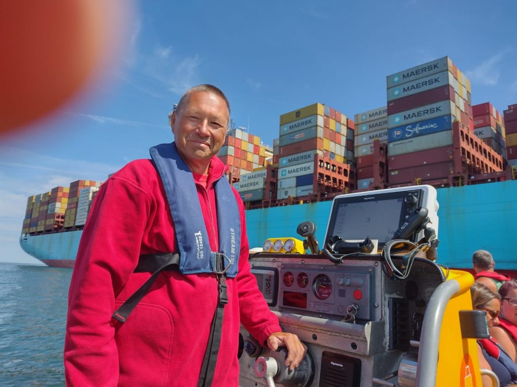 Skipper smiling at the helm of a jet boat next to a huge container ship