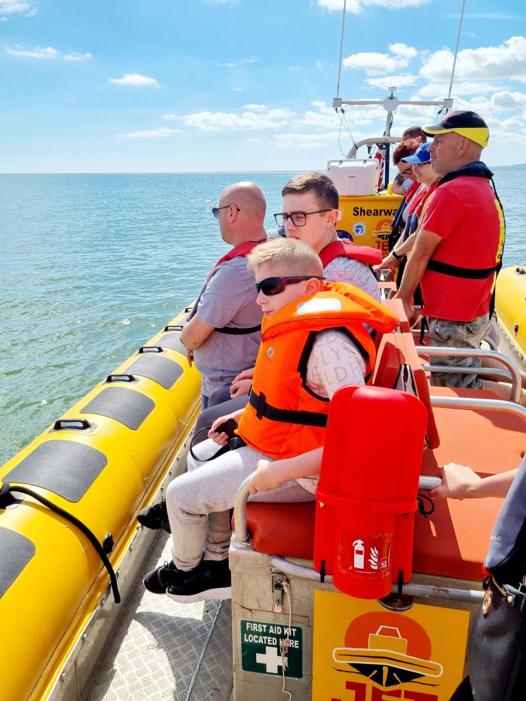 Group on rescue boat with life jackets, sunglasses, first aid kit; calm sea under clear sky.