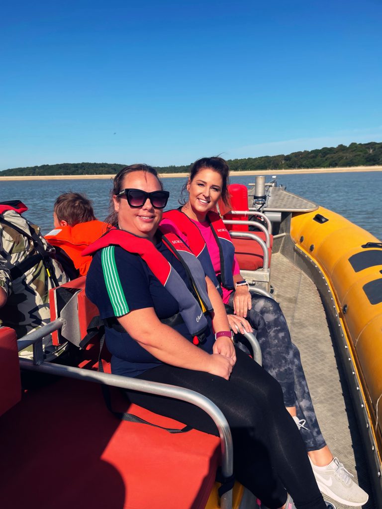 Two people on a boat in life jackets on a sunny day with a tree-lined shore in the background.